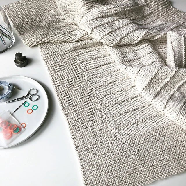 The On The Porch Baby Blanket Pattern by Fifty Four Ten Studio (Ravelry Instore) is shown in cream, with textured stripes. Scissors, stitch markers, and a tape measure suggest this easy pattern’s perfect for a cozy modern baby blanket in progress.