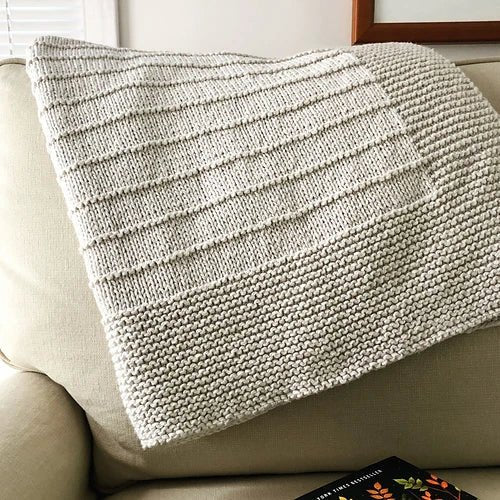 The "On The Porch Baby Blanket Pattern" by Fifty Four Ten Studio (Ravelry Instore, print) combines garter and stockinette stitch for a cream-colored knitted blanket, shown draped on a light beige sofa beside a floral-covered book.