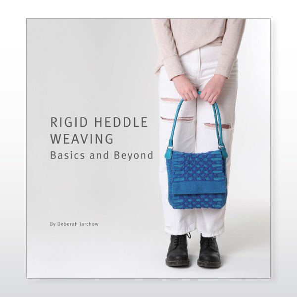 A person in a light sweater, ripped white jeans, and black boots holds a blue woven bag and "Rigid Heddle Weaving Basics And Beyond" by Deborah Jarchow from Ashford Handicrafts Limited.