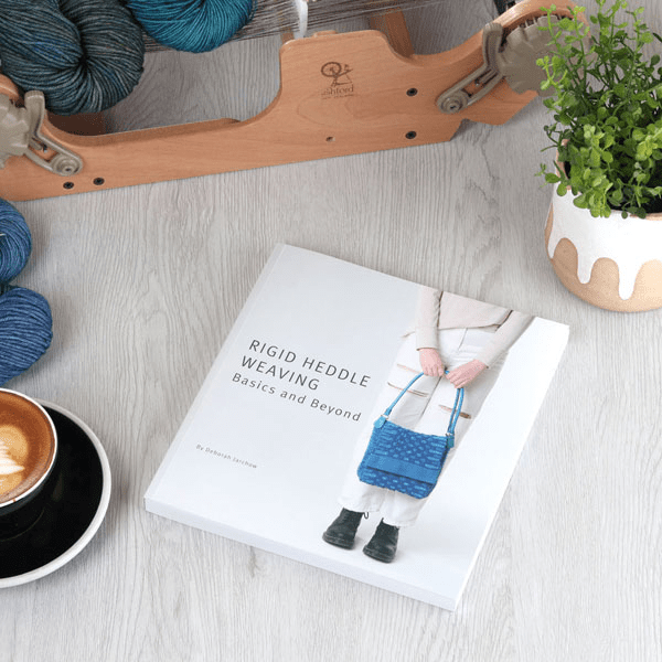 Rigid Heddle Weaving Basics and Beyond by Deborah Jarchow (Ashford Handicrafts Limited) sits on a light wood surface next to a cup of coffee, green plant, blue yarn, and an Ashford loom.