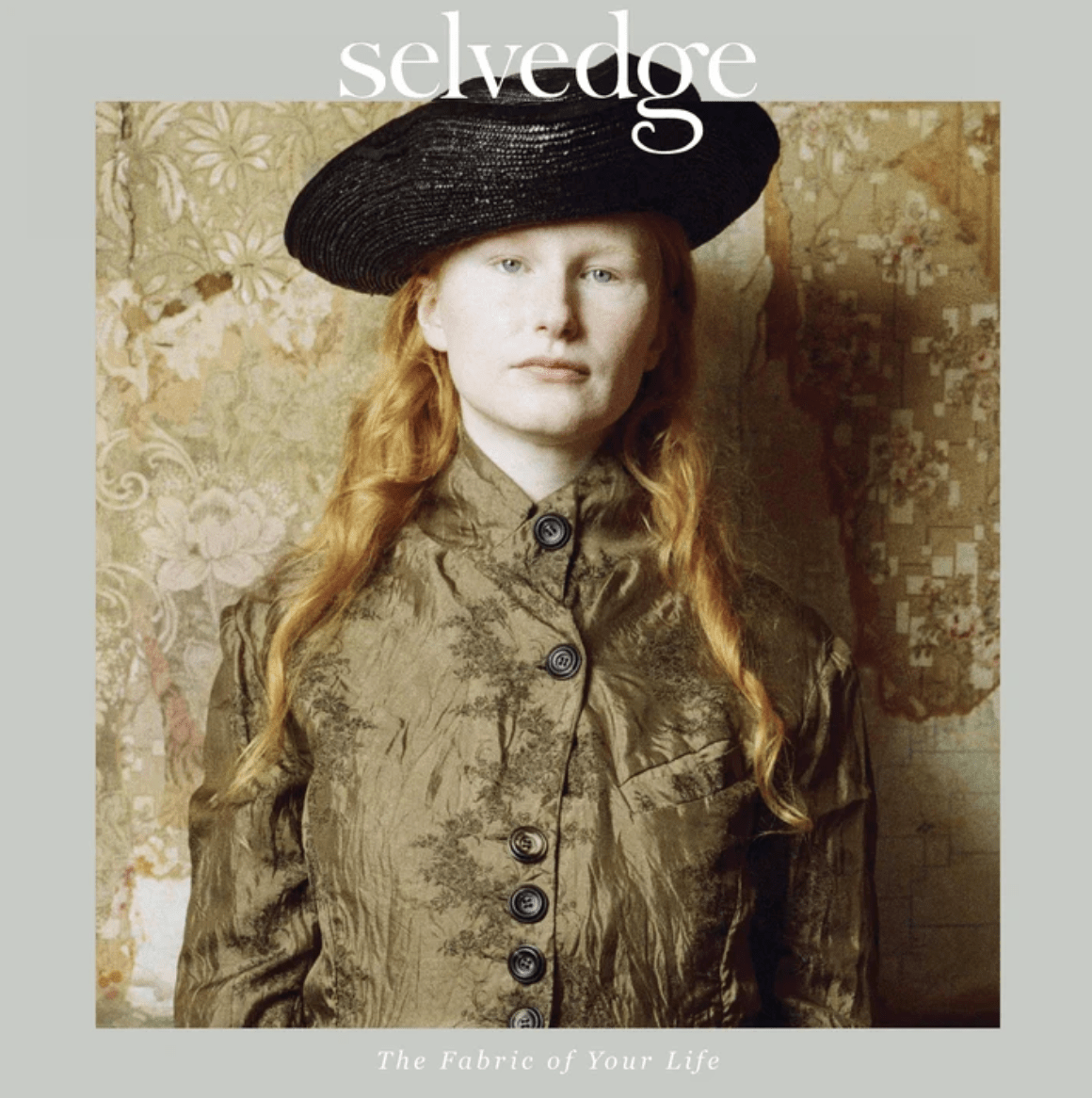 A person with long red hair wears a black hat and a brown buttoned jacket, standing against vintage floral wallpaper evocative of museum textile patterns. “Selvedge”—the brand name—appears at the top, while “Issue 132: Legacy” is noted at the bottom, conjuring connections to historic textiles such as the Bayeux Tapestry.