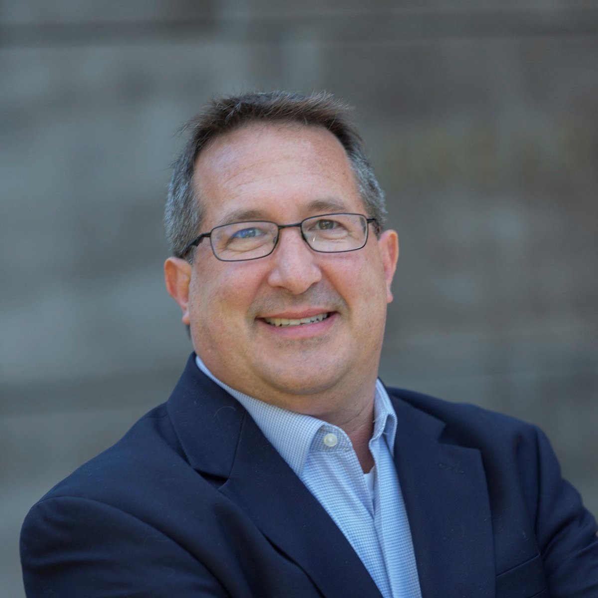 A man with short brown hair and glasses smiles at the camera in a navy blazer and light blue shirt, standing before a blurred gray background at the Halcyon Yarn Weaving Demo + Pop-Up Shop, Oasis Free Clinic Benefit, May 8–9.