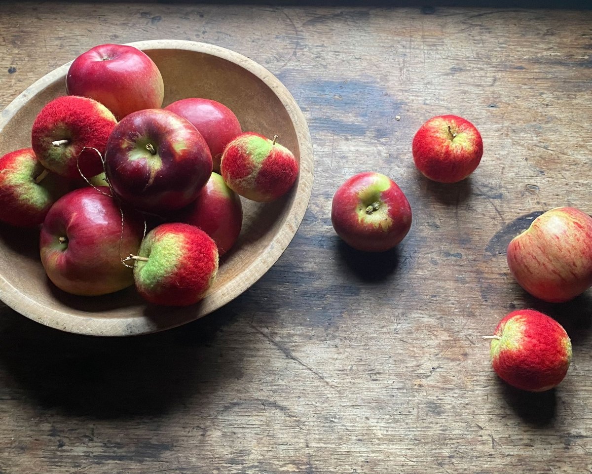 A rustic wooden table hosts a mix of red and green apples overflowing from a wooden bowl, while several lifelike felted wool apples—created through needle felting—are artfully arranged beside the bowl. The scene is softly illuminated from the top left, highlighting the realistic felted fruit featured in Halcyon Yarn’s Needle Felting Class: Apples with Michelle Provençal (September 12th).