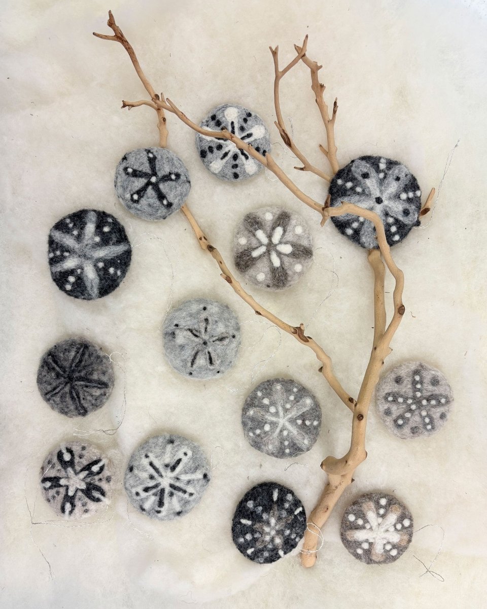 Fourteen round felted pieces, each crafted to resemble delicate sand dollars in shades of black, gray, and white, are arranged on a soft white surface with a bare light-colored branch placed diagonally across them—showcasing the beautiful results you can create in Halcyon Yarn’s “Needle Felting Class: Sand Dollars with Michelle Provençal” (September 5th).