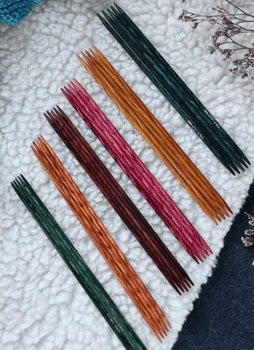 Dreamz Knit Pro 8" Double Point Knitting Needles by Accessories Unlimited, in green, orange, maroon, and pink birchwood, are arranged on textured white fabric with dried flowers—adding vibrant charm to any crafting space.