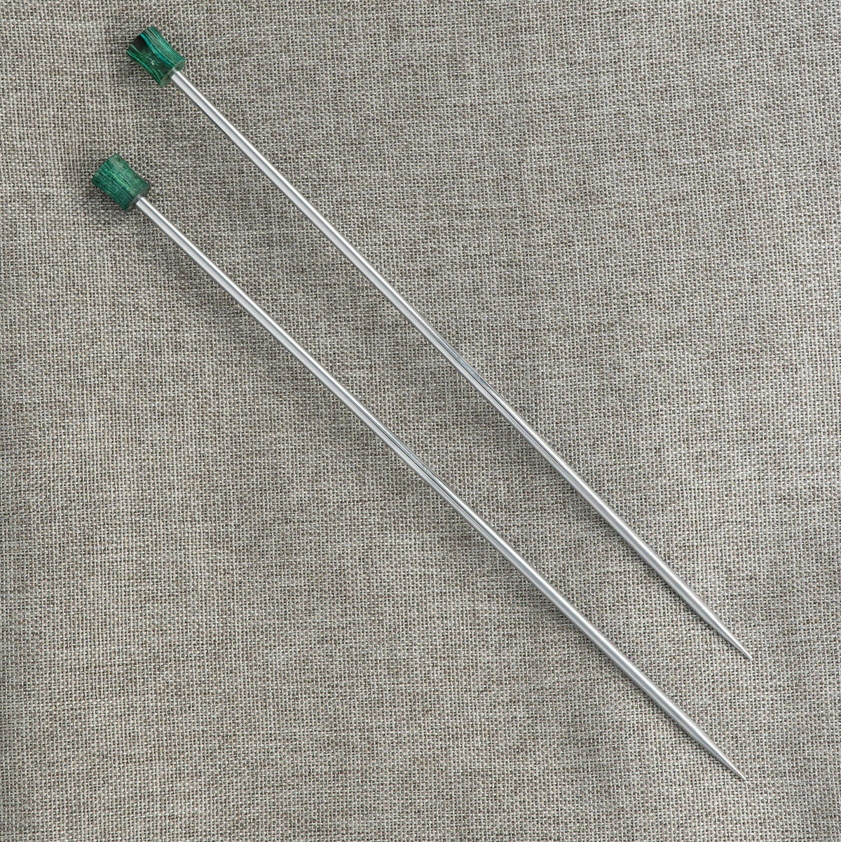 Two long Knit Pro (Knitters Pride) Nova Metal Single Point Knitting Needles by Accessories Unlimited are placed parallel on a textured gray fabric background, each featuring green handles and available in 10" or 14" lengths.