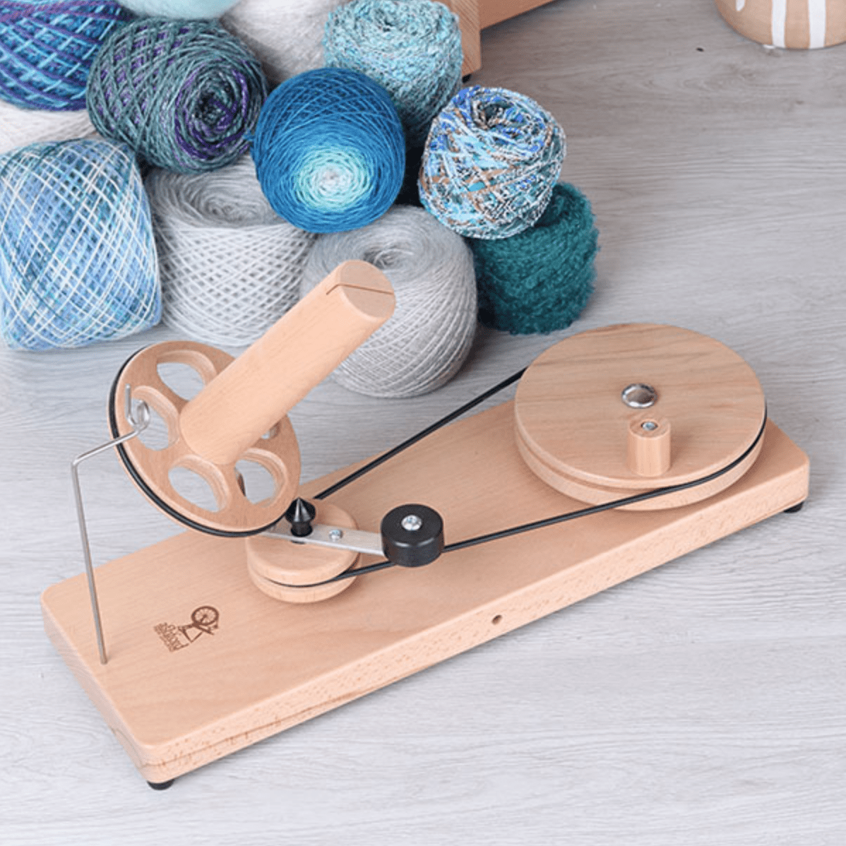 An Ashford Wooden Ball Winder by Ashford Handicrafts Limited, with a metal arm and handle, sits on a light floor. Behind it are several colorful yarn balls in blue, turquoise, and purple shades.