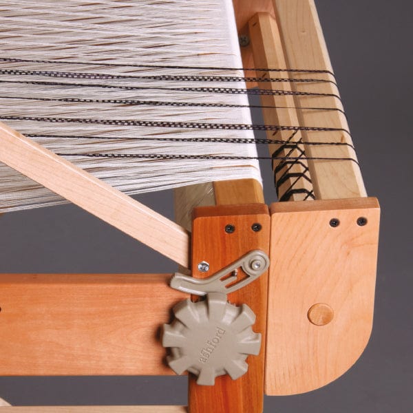 A close-up of an Ashford Second Back Beam for Table Looms by Ashford Handicrafts Limited shows white threads stretched across the frame, with a gray adjustment knob and darker threads interwoven among the lighter ones. The second back beam is partially visible, highlighting the intricate details of this weaving setup.