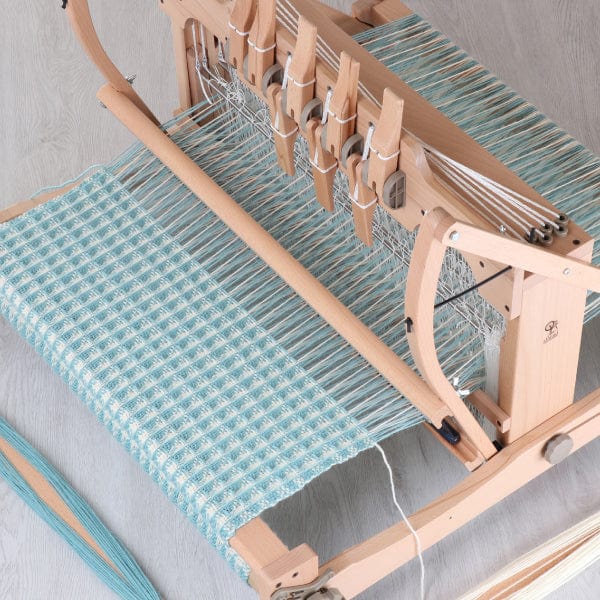 An Ashford Handicrafts Limited Table Loom with Overhead Swinging Beater and Shuttle Race, threaded in light blue and white for a checkered fabric, is placed on a light wood floor with threads and woven material extending from the loom.