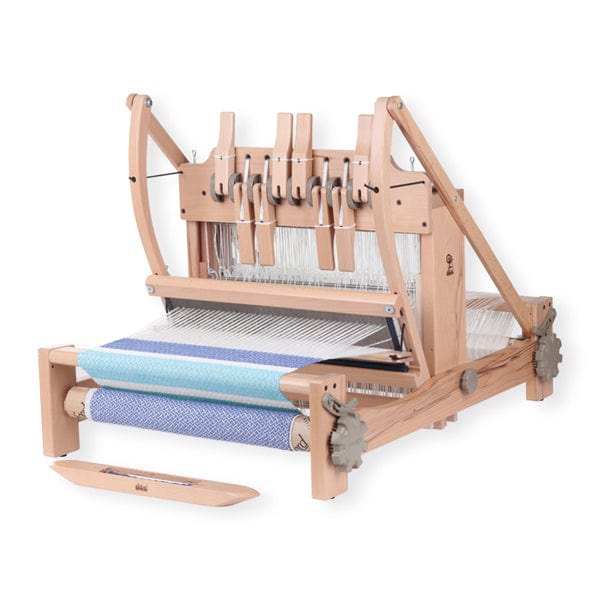 The Ashford Table Loom, by Ashford Handicrafts Limited, with overhead swinging beater and shuttle race, is weaving light blue and white patterned fabric—an ideal 8-shaft loom for intricate projects.