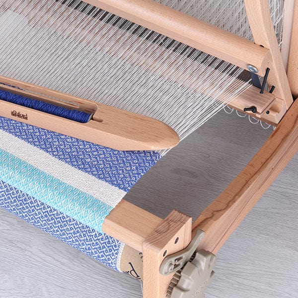 Close-up of Ashford Handicrafts Limited’s Ashford Table Looms with Overhead Swinging Beater and Shuttle Race, featuring blue, turquoise, and white threads woven into patterned fabric; unwoven white warp threads are tightly stretched.