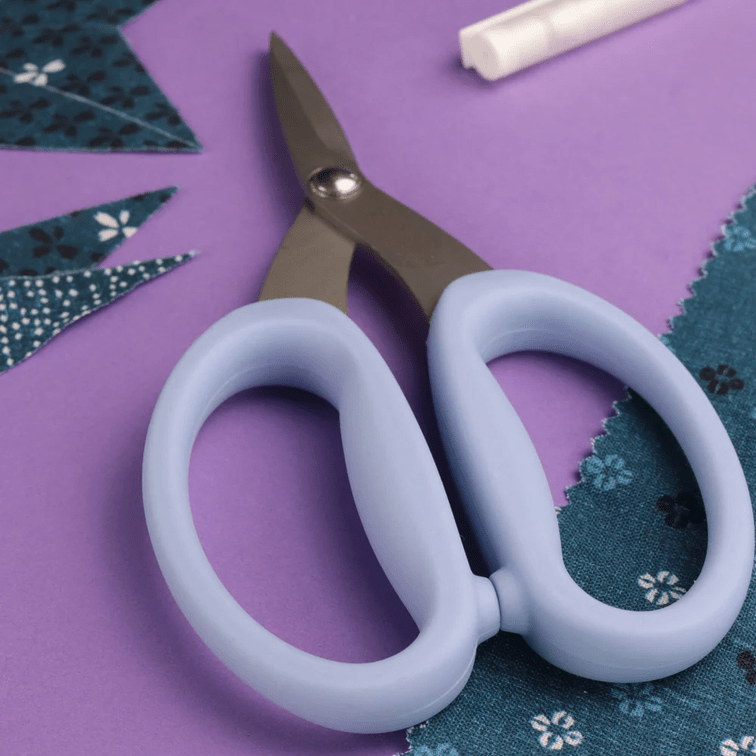 A pair of 6.75" Micro-Serated High-Precision Scissors by Bryson Distributing, Inc. with light blue handles lies on a purple surface near blue patterned fabric pieces and a partially visible white pen in the background.