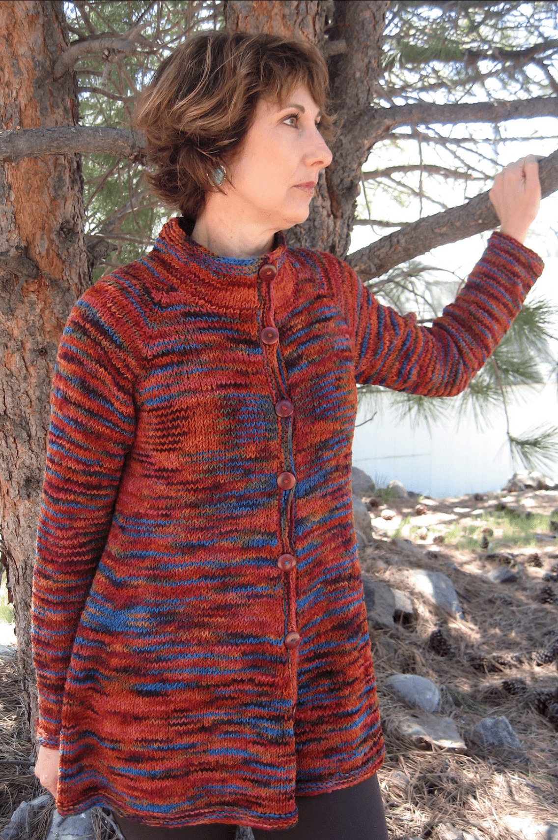 A woman stands outdoors by a pine tree, wearing the Knitting Pure & Simple Neck Down Swing Coat #285, a long, multicolored cardigan knit in worsted weight yarn with large front buttons. She looks to the side and touches a tree branch.