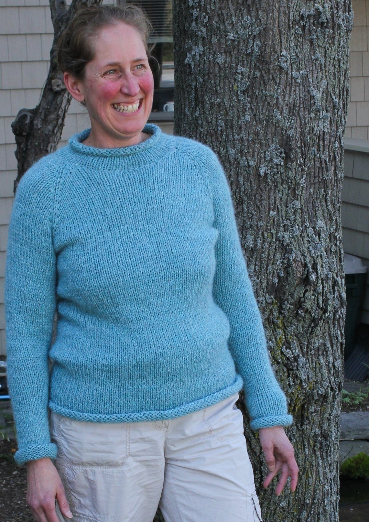 A smiling person stands outdoors beside a tree, with a building in the background, wearing the Knitting Pure & Simple Bulky Neck Down Pullover #992 in light blue and beige pants.