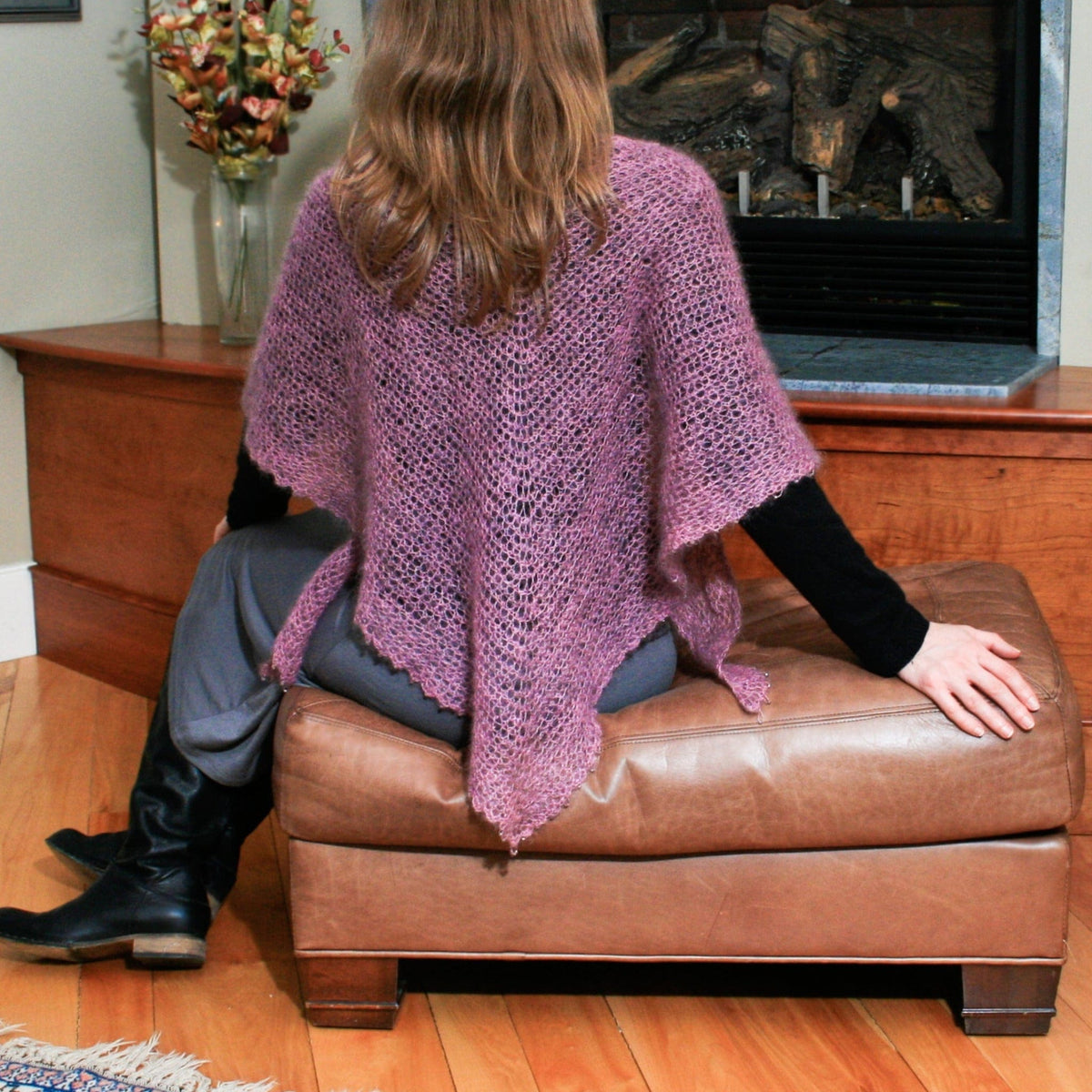 A person viewed from behind sits on a leather ottoman wearing a mauve, open-knit Crocheted Cobweb Capelets & Shawls, a design similar to a capelet pattern.