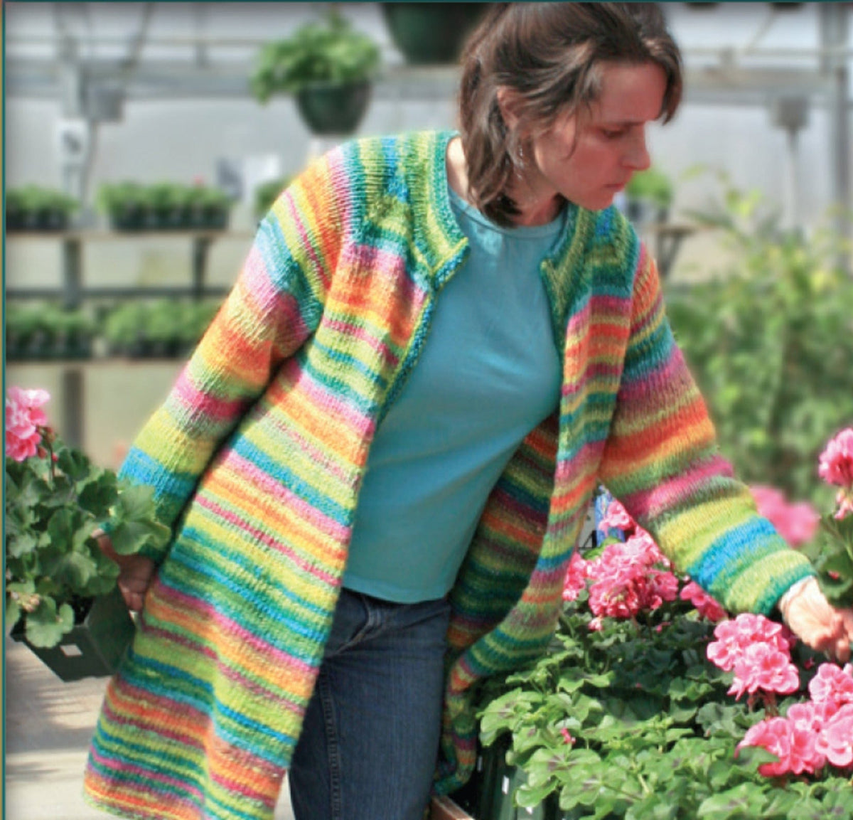 A woman in the Knitting Pure & Simple Neck Down Swing Coat (#285, download), a colorful striped cardigan made from worsted weight yarn, reaches toward pink flowers in a greenhouse filled with potted plants.