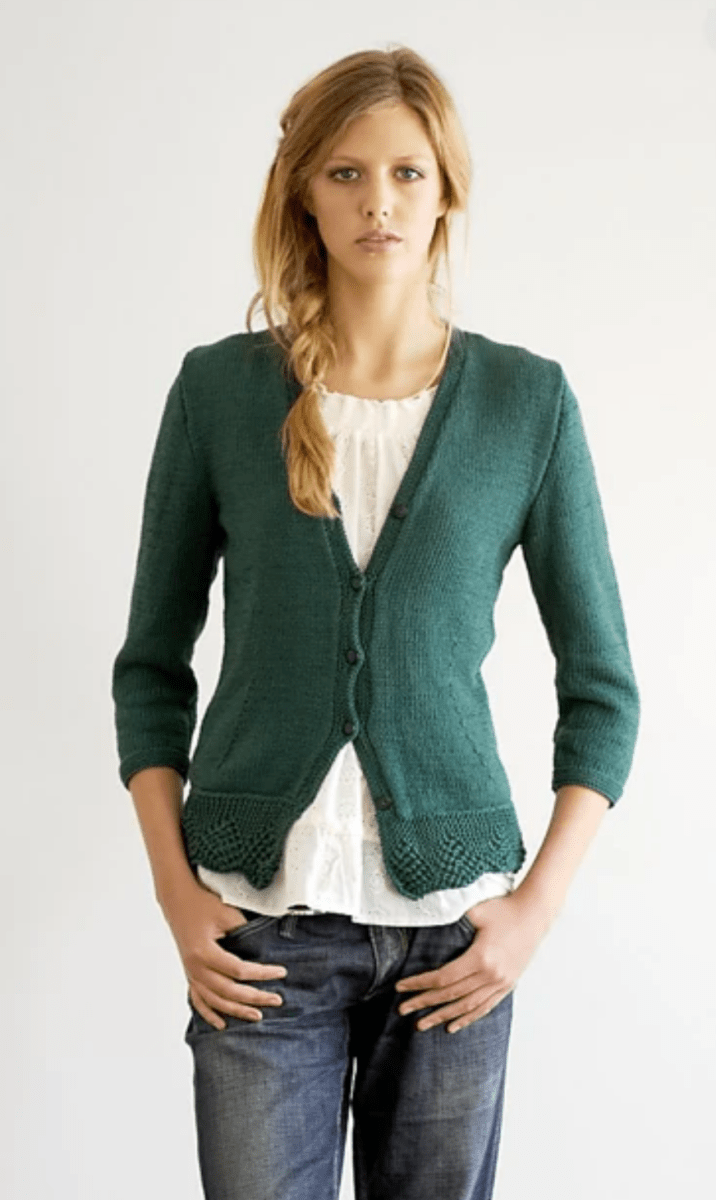 A woman wears a forest green Fitted Cardigan, by Jo Sharp, featuring a fitted, button-up design with three-quarter length sleeves and a decorative knit hem over a white blouse and blue jeans.