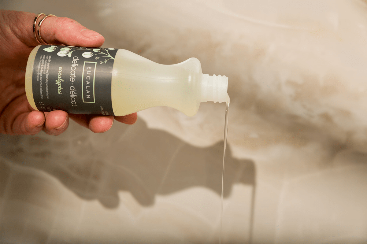 A hand pours liquid from a bottle labeled “Eucalan Wool Wash 100 ml / 3.3 oz” by Eucalan, a lanolin-enriched, biodegradable textile cleanser, against a light, blurry background.