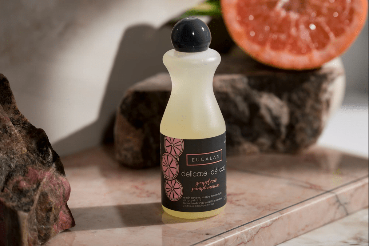 A 100 ml / 3.3 oz bottle of Eucalan Wool Wash by Eucalan in grapefruit scent rests on a marble surface, accompanied by a sliced grapefruit and rocks in the background. This biodegradable, no-rinse textile cleanser is perfect for gently washing wool and other delicate fabrics.