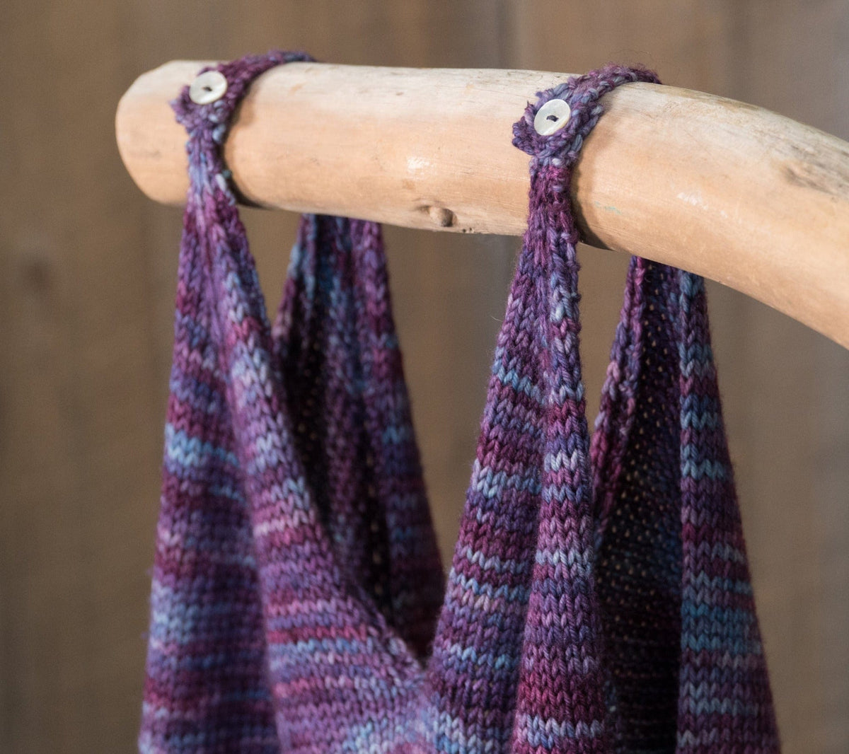 Close-up of the Flower In The Sun Tank Top by Sierra Roberts from Halcyon Yarn, featuring sport weight yarn in purple, blue, and gray hues, displayed on a wooden rod with straps secured by white buttons. Downloadable pattern.