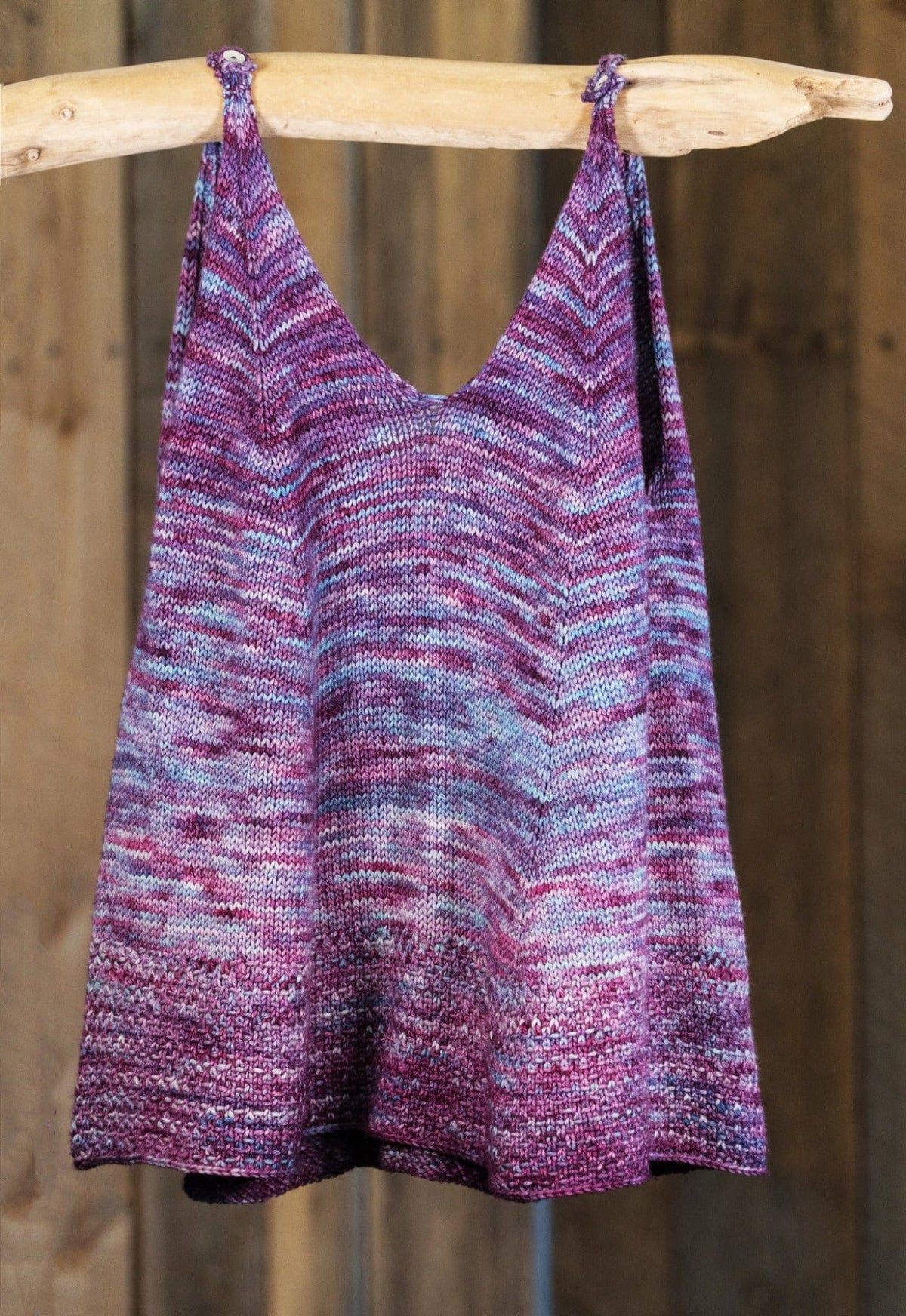 The Flower In The Sun Tank Top by Sierra Roberts, from Halcyon Yarn, is a sleeveless knit tank in purple and gray sport weight yarn, shown hanging from a wooden rod against a rustic wood background. Available as a digital download.