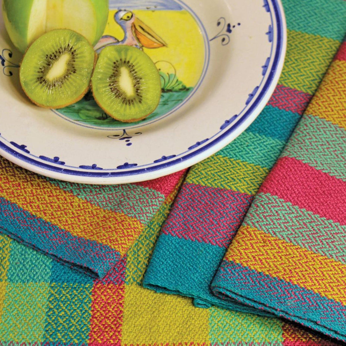 A plate with a pelican illustration holds a green apple half and kiwi atop Halcyon Yarn's Eight Organic Cottolin Towels, featuring bright yellow, pink, turquoise, and orange handwoven patterns. Download available.