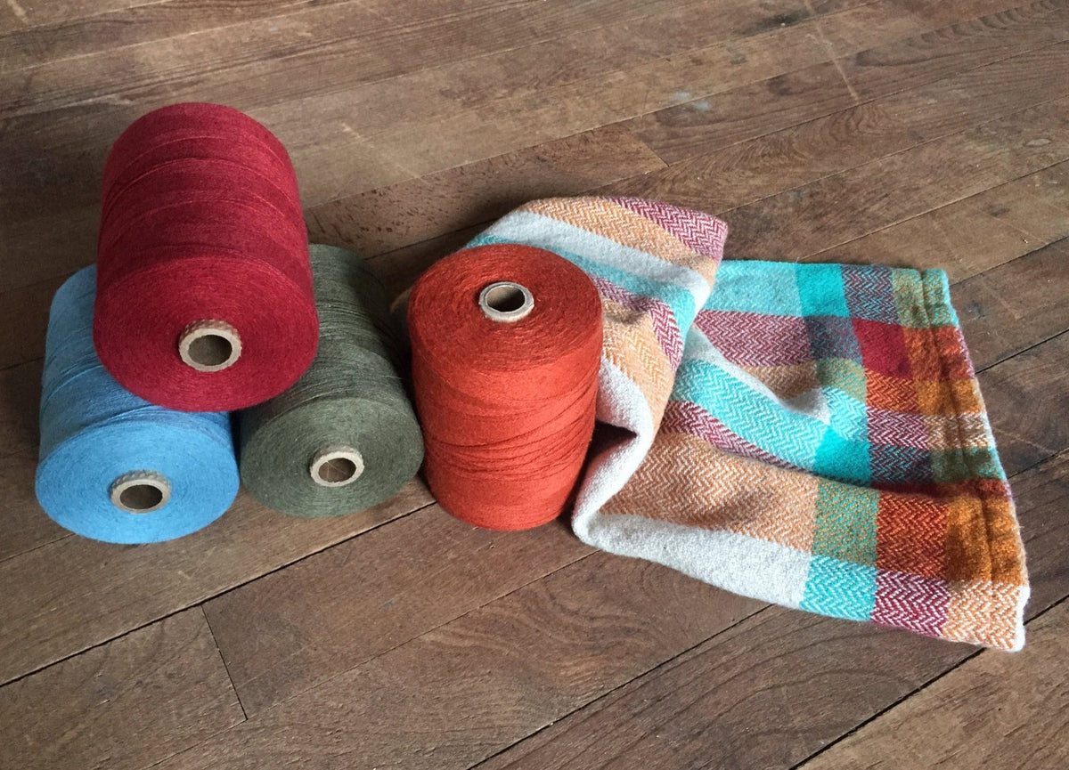 Four large yarn spools in red, blue, green, and orange rest on wood beside a folded Halcyon Yarn Eight Organic Cottolin Towels download set, handwoven in orange, teal, yellow, and beige from cottolin on a 4-shaft loom.