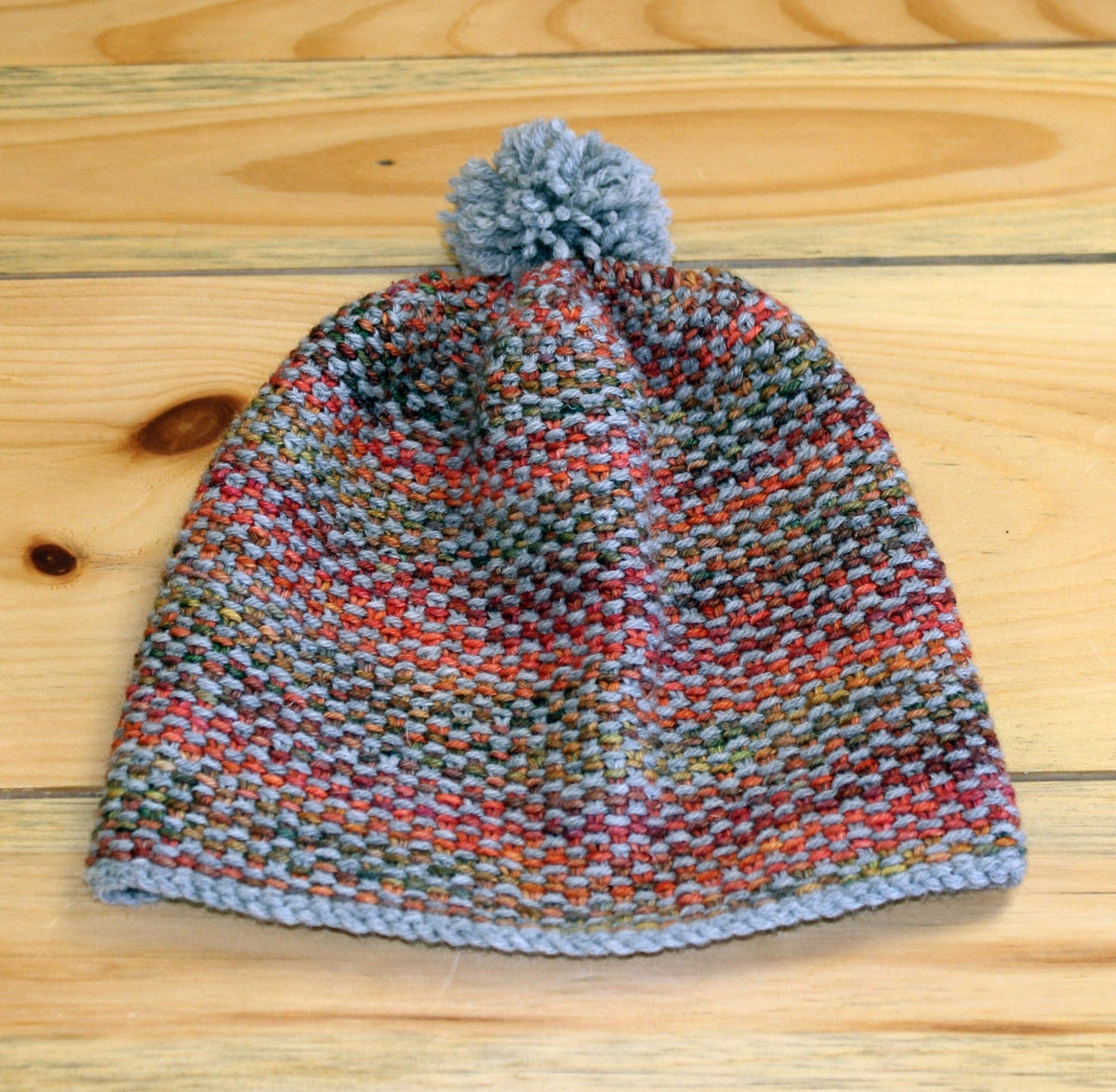 The Telephone Line Hat & Cowl by Halcyon Yarn is a slip-stitch knit beanie in multicolored red, green, and orange tones with a light gray edge and pom-pom, crafted in Jo Sharp Classic DK Wool and shown on a wooden surface. Download available.