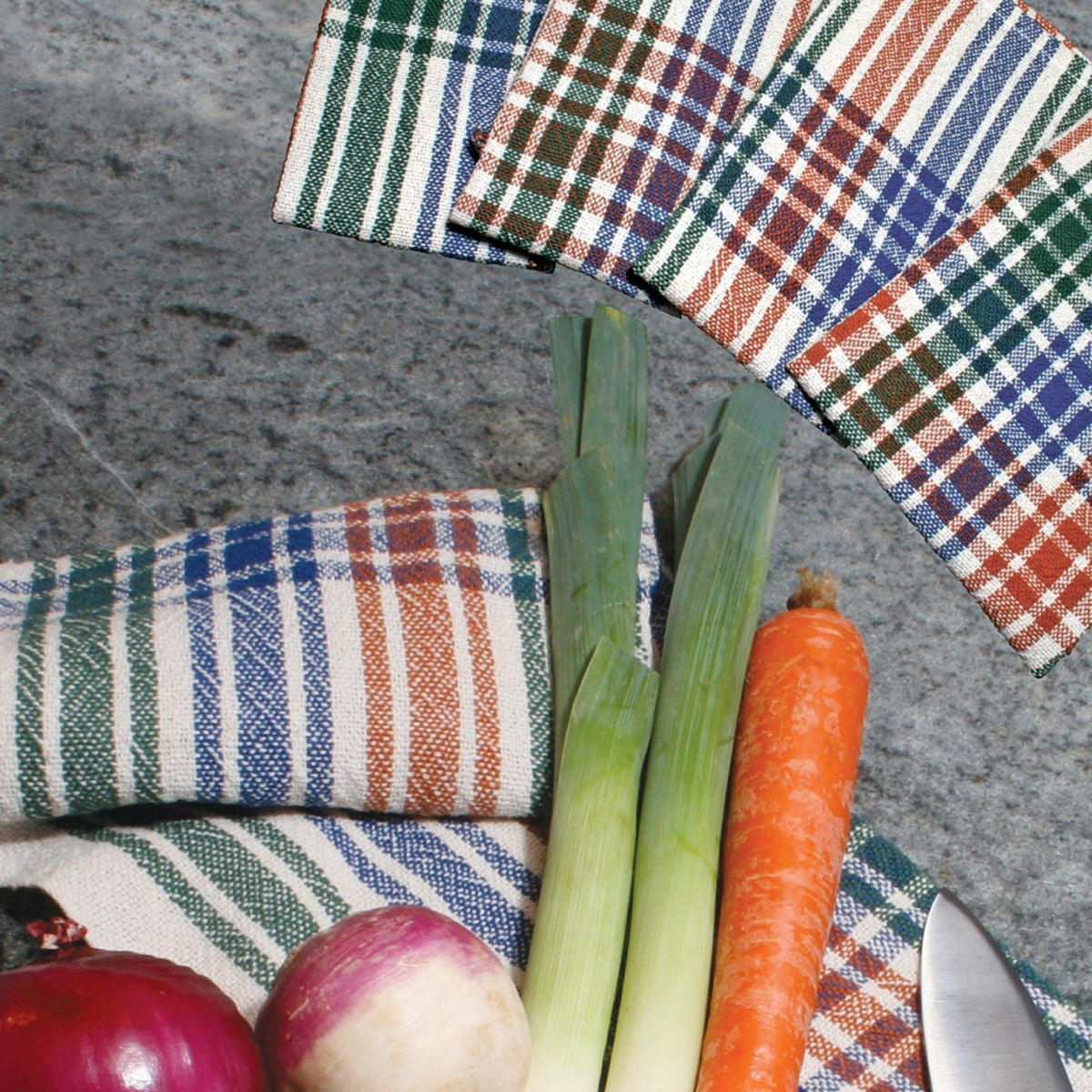 A red onion, turnip, two leeks, and a carrot rest on plaid cloth woven using Halcyon Yarn’s Camp & Cottage Towels Weaving Pattern (4-shaft & rigid heddle), with a kitchen knife and matching napkins on a gray stone surface.