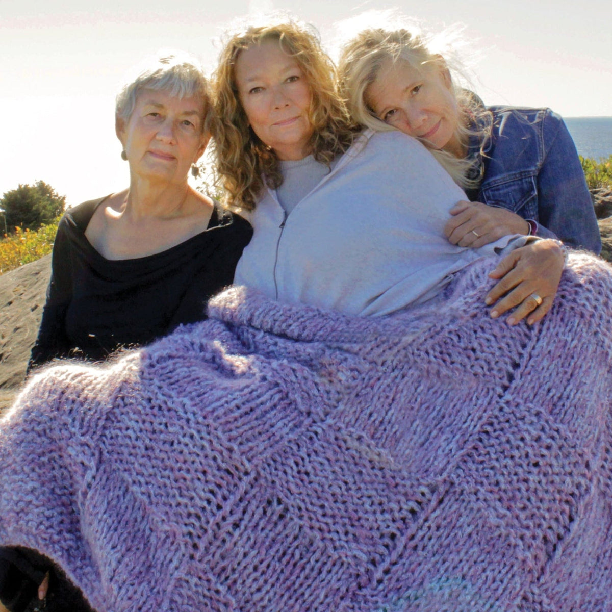 Three women sit closely together outdoors, wrapped in a Cobblestone Cove Victorian Throw by Halcyon Yarn. They smile softly with trees and the ocean in the sunny background.