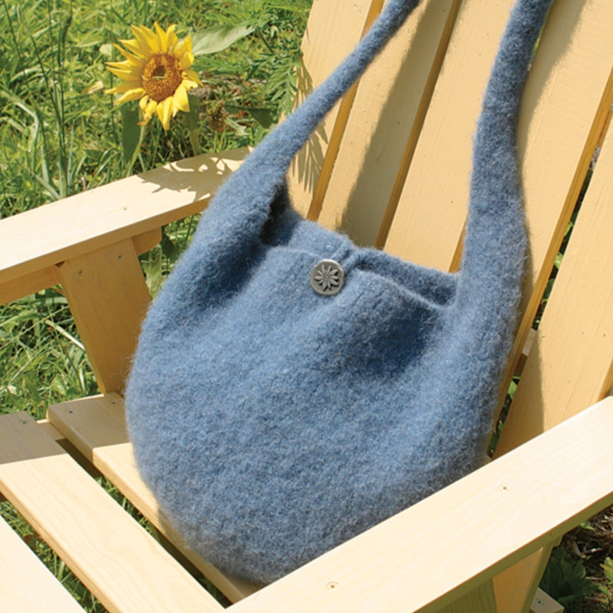 A Halcyon Yarn Felted Satchel Knitted in soft blue wool with a decorative button sits on a light wooden outdoor chair, with a sunflower blooming in the grassy background.