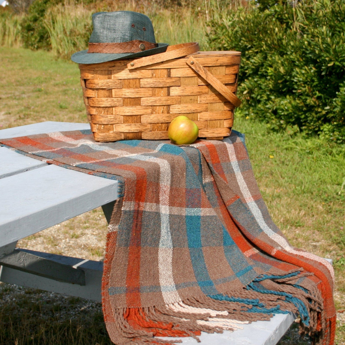 The Halcyon Yarn Fall Picnic Blanket Woven is displayed on a white wooden picnic table outdoors with grass and bushes behind, alongside a woven basket, green hat, and apple. The basket shows an intricate weaving pattern.