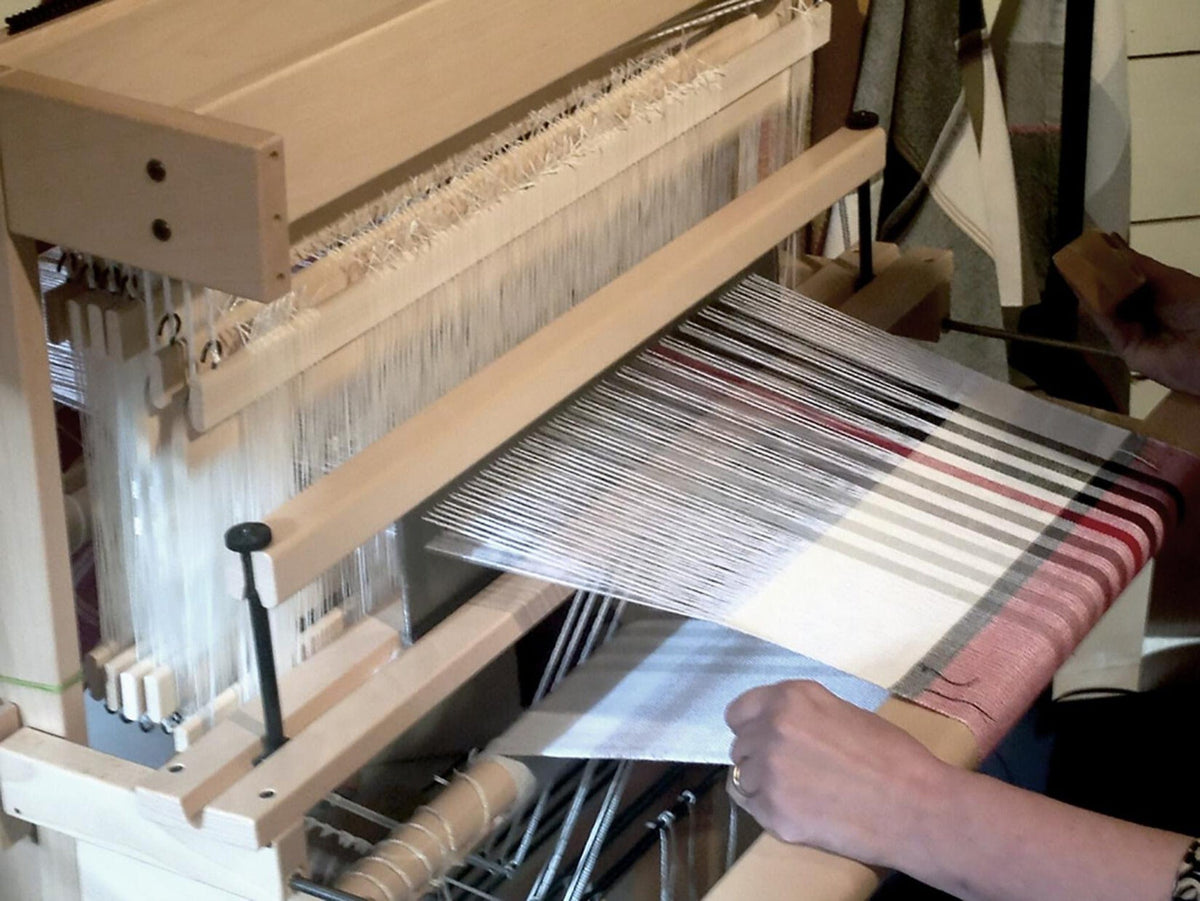 Using the Louët Sliding Beater for David Looms, a weaver skillfully guides threads into a fabric with white, black, and red stripes. Their hands deftly maneuver on visible wooden components to craft vibrant patterns.