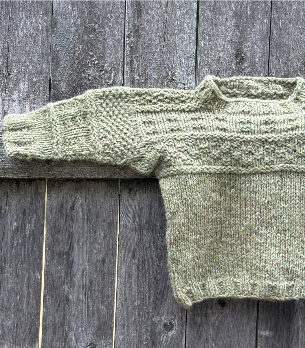 A hand-knitted mini "Everything Gansey" sweater in light green, created with Halcyon Yarn and designed for children, featuring long sleeves. The partially finished British gansey-inspired sweater is shown flat on a weathered wooden surface, displaying only one sleeve and half of the garment. Based on "Knit Your Own Mini Sweater with Linda Perry" (October 3, 2026).