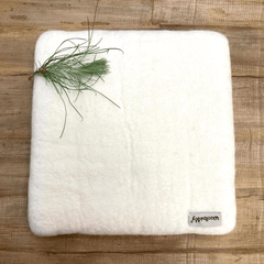 A square, white Woolbuddy Needle Felting Mat Large by Woolbuddy sits on a wooden surface with a small green pine branch at the top left and a "woodwoow" tag attached to the bottom right corner.