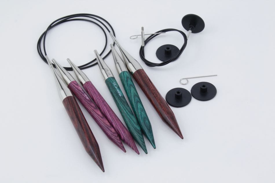 The Accessories Unlimited Knit Pro Dreamz Interchangeable Circular Knitting Needle Set features five colorful needle tips with metal ends, two black cables, end caps, and cable keys—ideal for chunky knitting—arranged on a white surface.