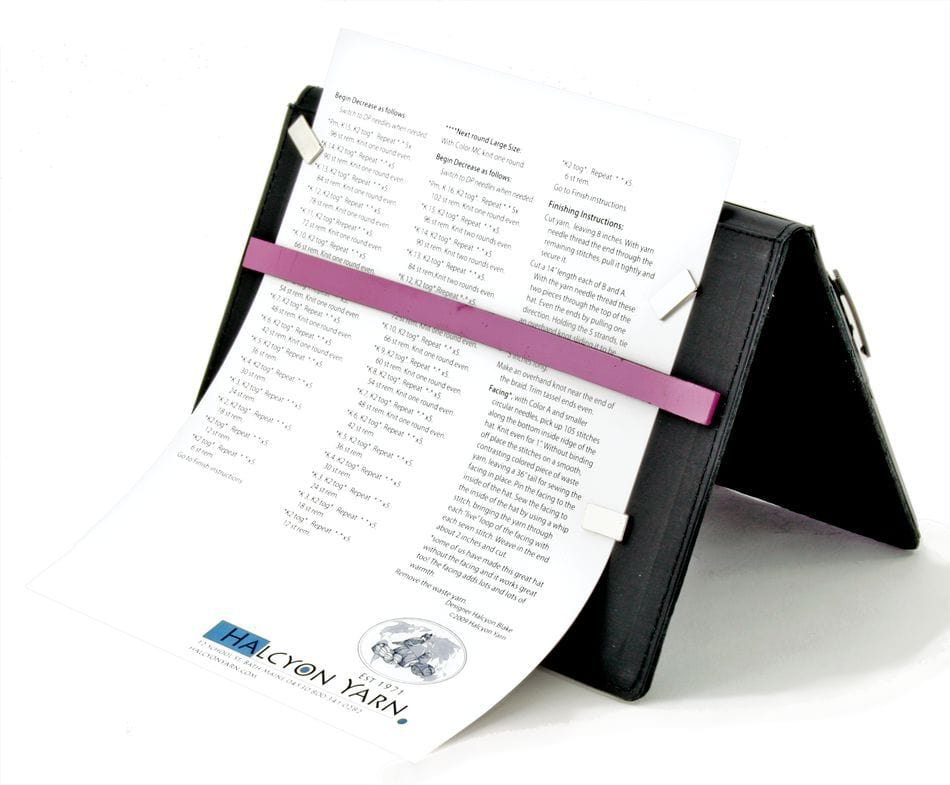 The Magnetic Pattern Holder by Knit Pro (Knitters Pride), from Accessories Unlimited, is a black, foldable stand with clips and a pink bar to hold your printed sheet upright, ideal for displaying patterns such as those featuring the Halcyon Yarn logo.